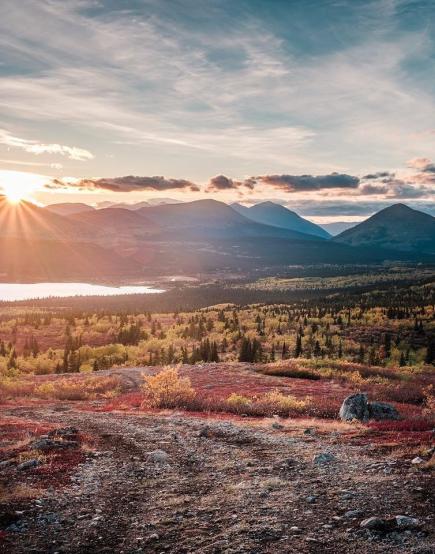 The sun sets over tall mountains at the base of a lake surrounded by the reds and oranges of fall